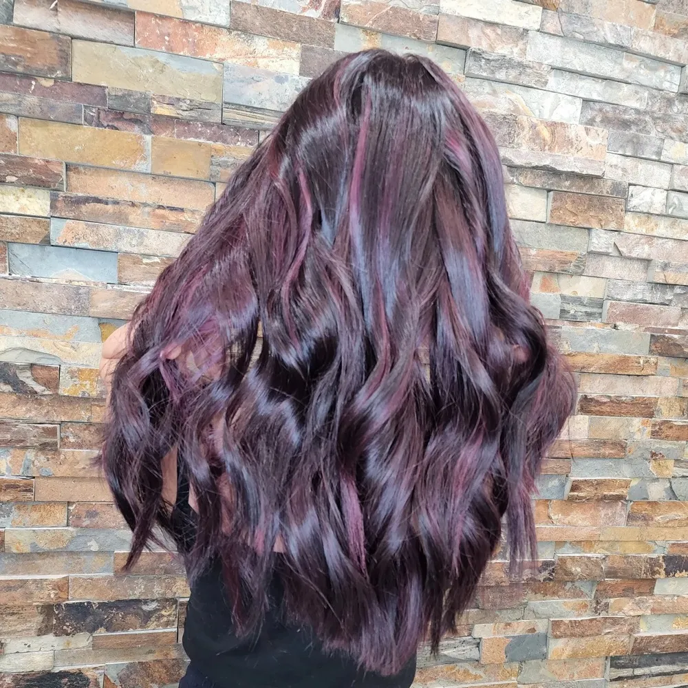 Long wavy hair colored in deep plum and violet, seen from behind against a stone wall